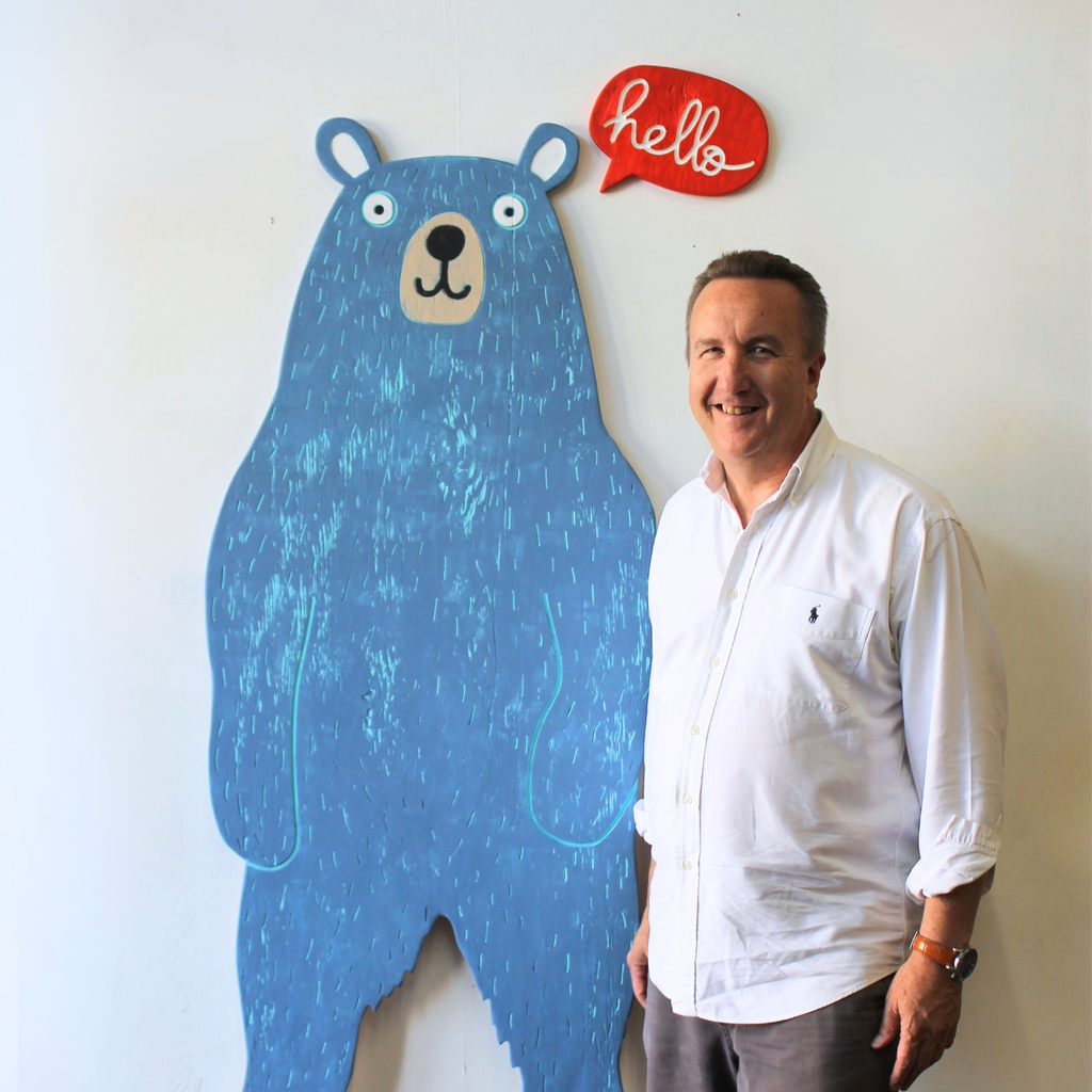 A smiling man (Gregory Morris-Artist) in a white button-down shirt stands next to a large blue bear-shaped wall decoration with a red speech bubble that says 'hello' on a white wall.