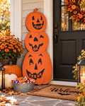 A outdoor three-tiered orange wooden jack-o'-lantern stack with carved faces and the words 'TRICK OR TREAT' on the front, displayed on a front porch next to a black door, a 'Welcome' doormat, a metal bucket of candy, and fall flowers.